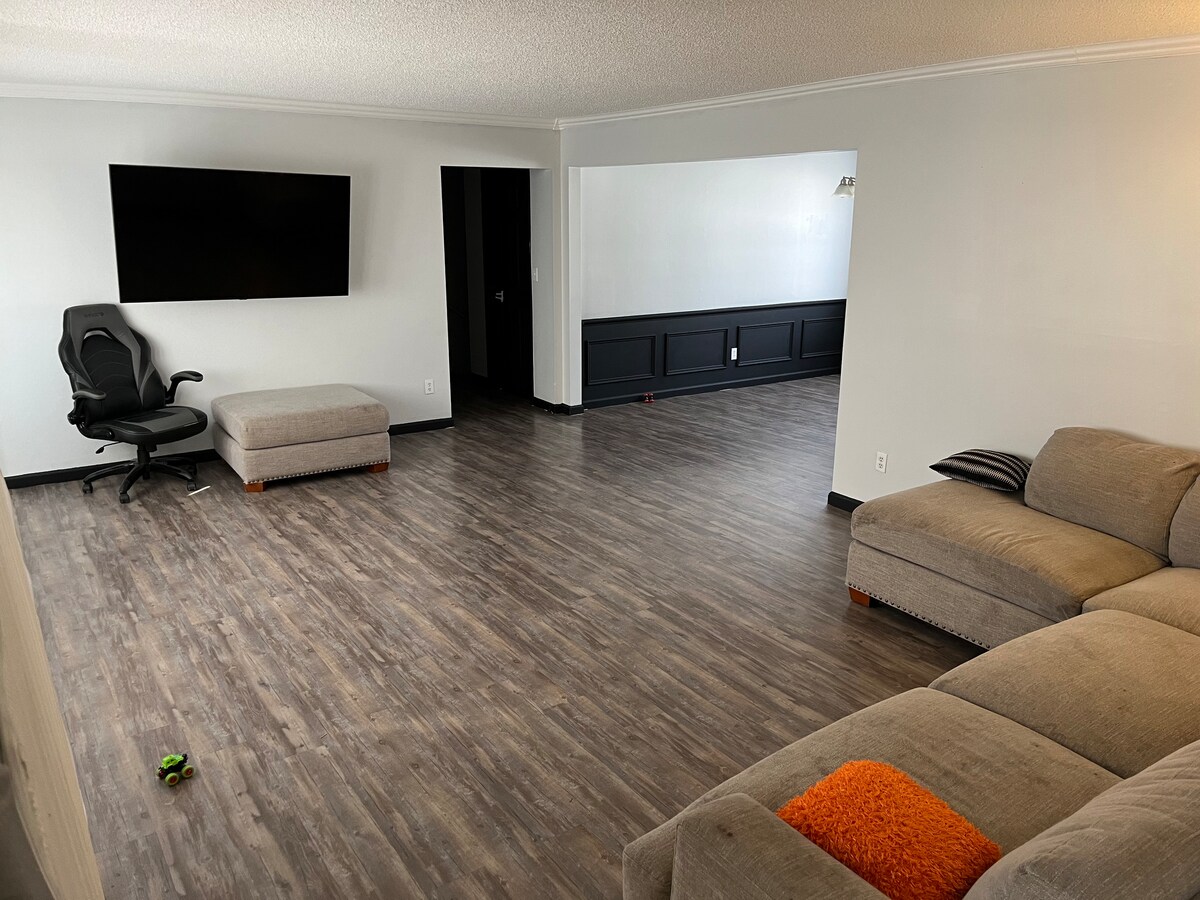A spacious living area is highlighted by a large flat-screen TV mounted on the wall. A sectional couch and an ottoman provide comfortable seating. The room features laminate flooring with a clean, modern aesthetic, and a wall with a dark finish adds character.