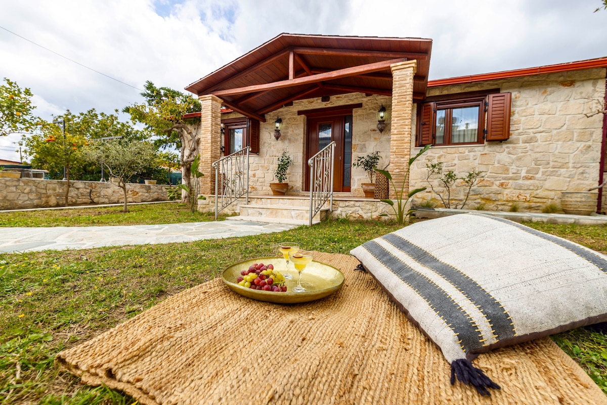 A charming stone house features a welcoming entrance with a covered porch. A basket of grapes and drinks is set on a woven mat in the lush garden, framed by vibrant greenery and decorative potted plants, inviting relaxation in a tranquil outdoor setting.