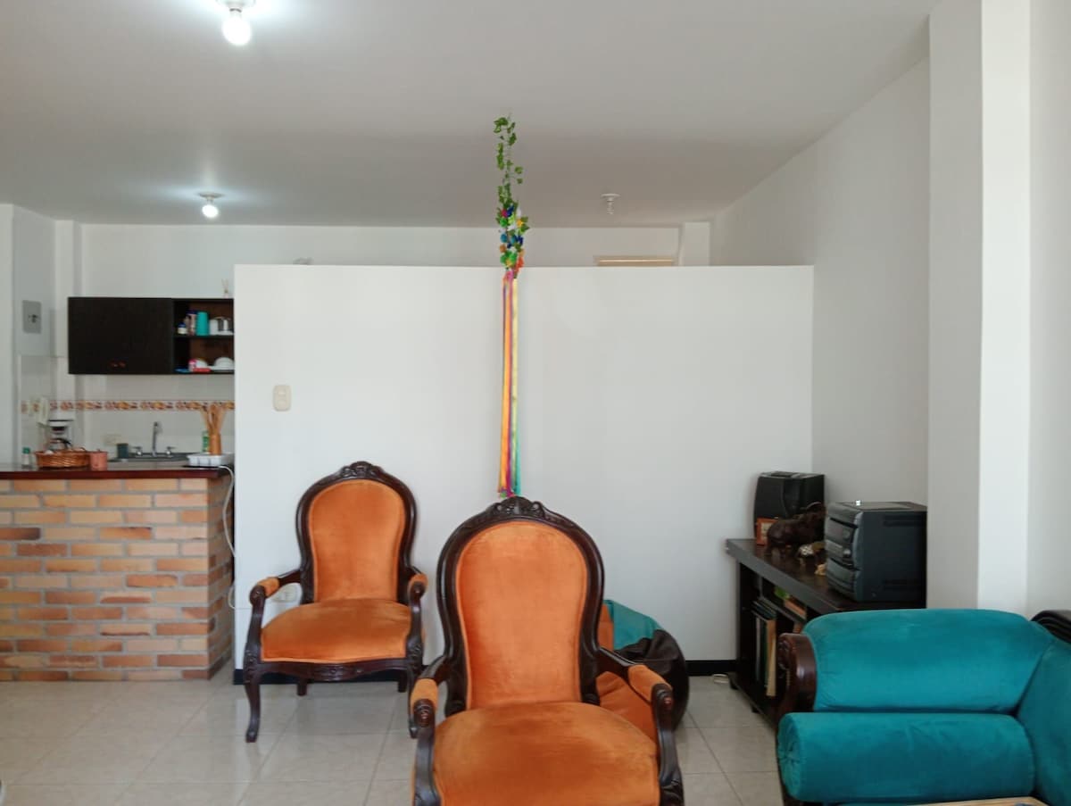A living area is presented with two orange armchairs facing a decorative division. A small kitchen is visible in the background, featuring wooden cabinets and various kitchen items. A teal couch complements the warm tones, and light is provided by overhead fixtures.