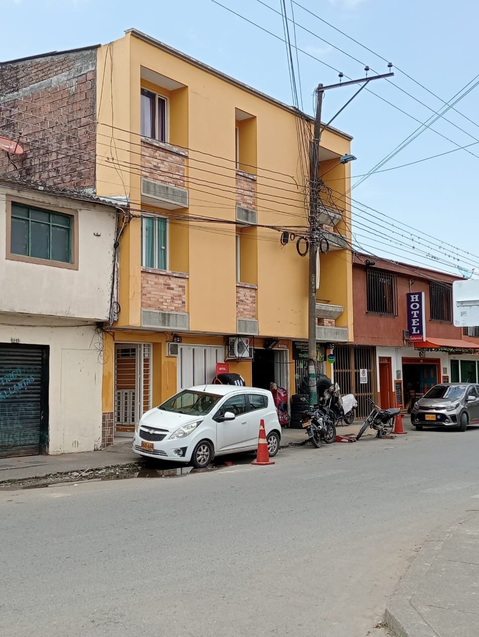 A three-story building is shown with a yellow facade and brick accents. The ground level features commercial spaces with a variety of signage. Parked cars and motorcycles line the street, while power lines intersect above, creating a lively urban scene.