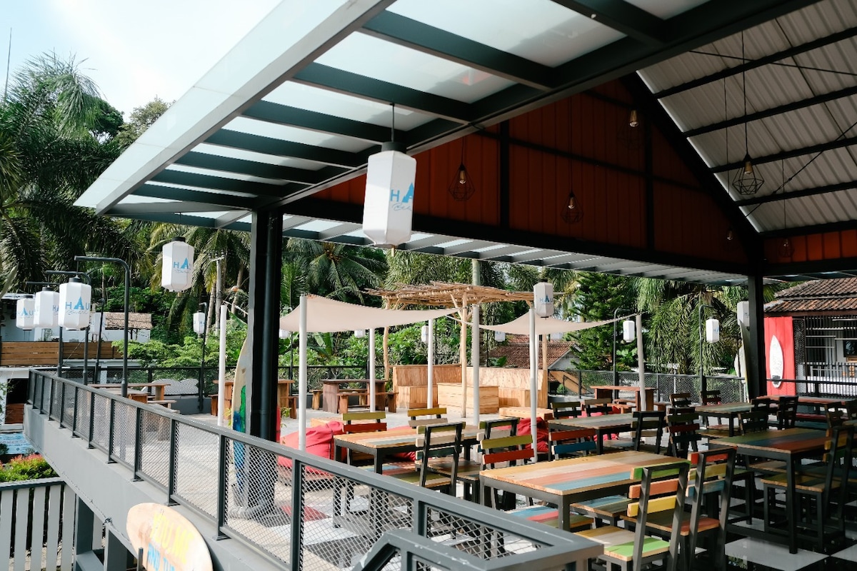 An outdoor dining area is revealed, featuring a series of wooden tables and chairs arranged under a covered structure. Lush greenery is visible in the background, providing a natural setting. Lanterns hang from the ceiling, enhancing the inviting atmosphere.