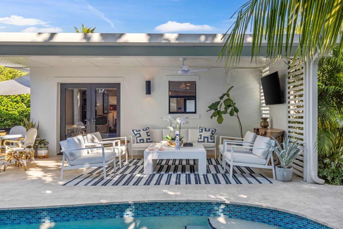 An outdoor seating area is displayed, featuring comfortable white chairs surrounding a central table atop a striped rug. Lush greenery is present, and a modern wall-mounted TV is visible. The private pool reflects the surrounding sunlit area, inviting relaxation.
