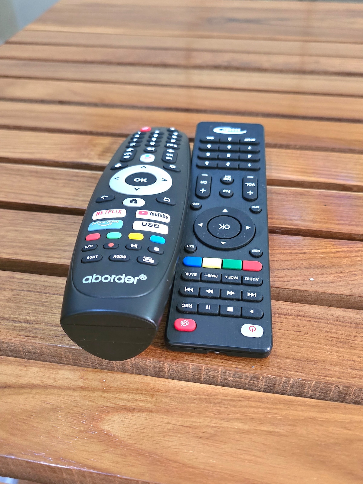 Two remote controls are placed on a wooden surface, showcasing various buttons for navigation and functionality. One remote is labeled 'aborder,' with a combination of red, green, yellow, and blue buttons, while the other features a different layout with standard playback controls.