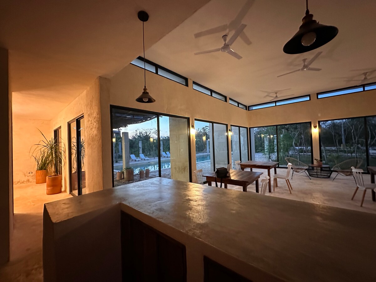 An open common area features large windows that bring in natural light, offering views of the surrounding greenery. The space includes a wooden bar counter and dining tables with comfortable seating, along with ceiling fans for airflow.