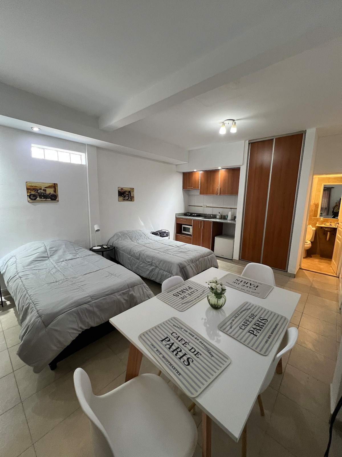 A spacious living area is arranged with two individual beds covered in gray bedding. A dining table with white chairs sits in the center, complemented by placemats. A kitchen area is visible in the background, featuring wooden cabinetry and essential appliances.