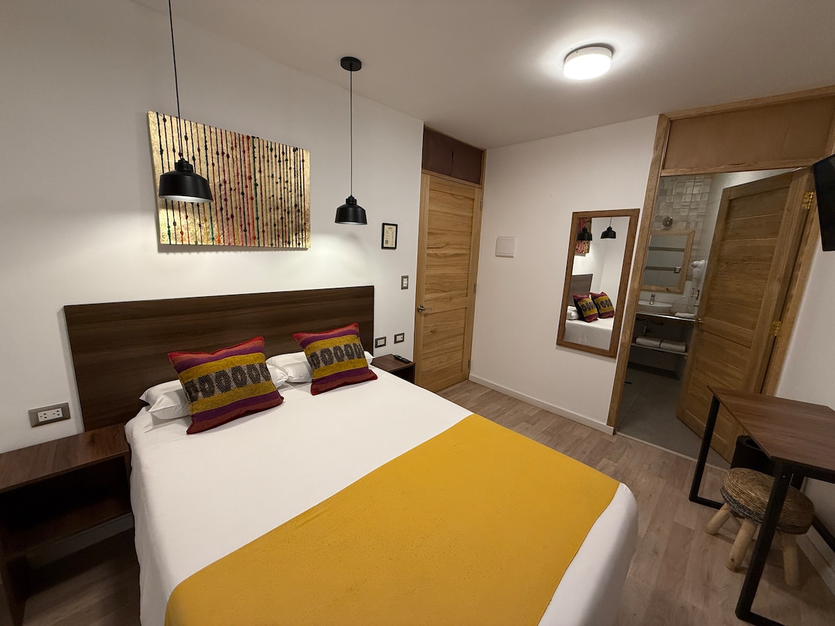 A well-appointed bedroom features a spacious bed with colorful textile accents. Soft lighting is provided by modern pendant lamps. A mirror reflects the room's neutral decor, complemented by wooden elements, while a door leads to the private bathroom.
