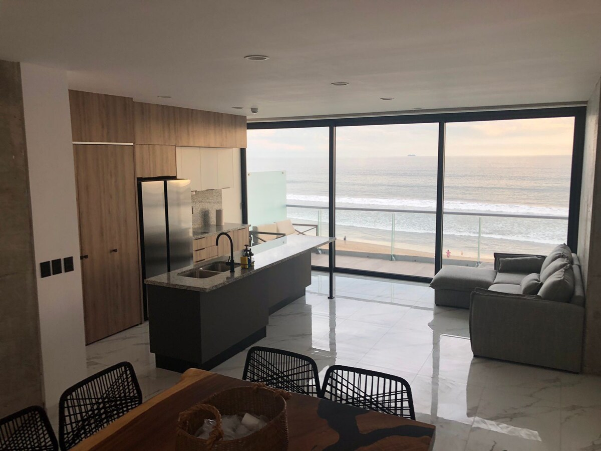 An open-concept living area features large glass doors that provide a view of the beach. A sleek kitchen with modern cabinetry is complemented by a light-colored countertop. A comfortable sofa is positioned in front of the ocean view, enhancing the sense of space and tranquility.