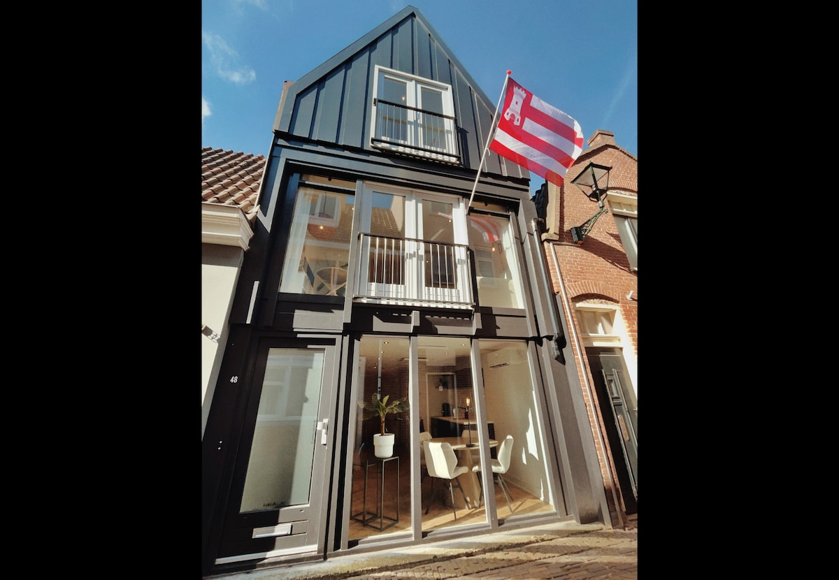 The exterior of the building features sleek black wood siding and large glass doors, inviting natural light into the space. A flag is displayed prominently above the entrance, and outdoor seating is visible on the balcony, enhancing the urban ambiance.