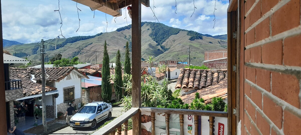 A view from a balcony showcases a panoramic landscape of rolling hills and distant mountains under a partly cloudy sky. The foreground includes rustic buildings with terracotta roofs, framed by greenery and small trees lining the street.