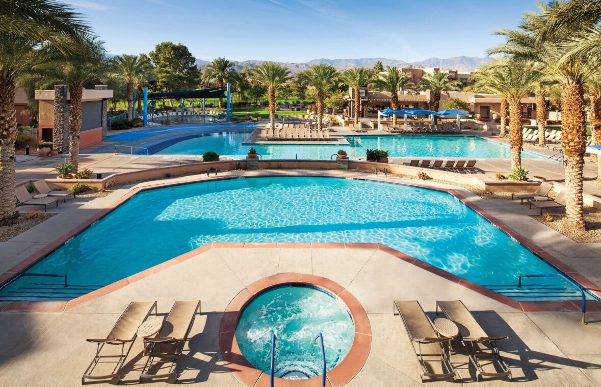 A vibrant outdoor pool area is surrounded by palm trees, featuring a large swimming pool and a separate hot tub. Multiple lounge chairs are arranged along the deck, offering a space for relaxation. The backdrop includes a clear blue sky and distant mountains.