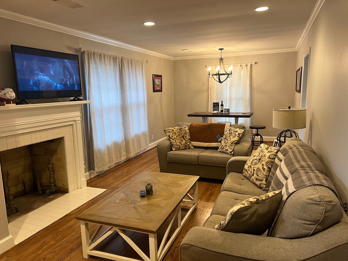 A living room is presented with two comfortable sofas adorned with patterned throw pillows. A coffee table is positioned in front of the seating area, while a traditional fireplace adds warmth to the space. Light-filtering curtains frame the windows, and a television is mounted above the mantel.