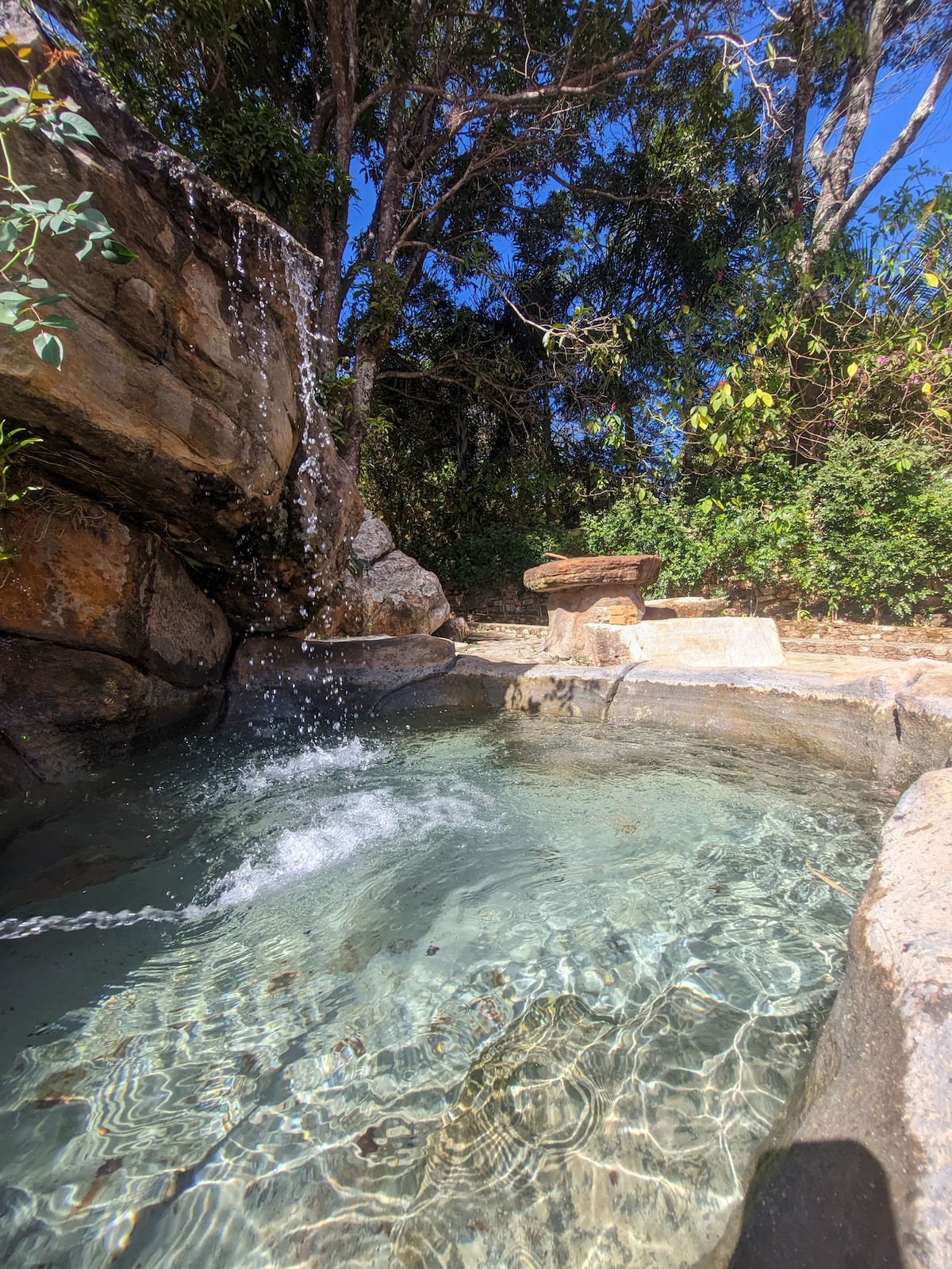 A natural stone hot tub is surrounded by lush greenery, with a gentle waterfall cascading over rocks into the clear water. Sunlight reflects off the surface, creating a serene ambiance. A smooth stone table is positioned nearby for relaxation.