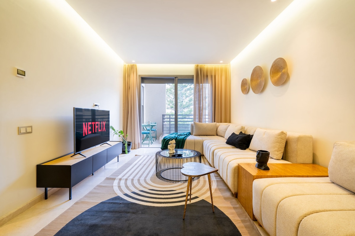 A modern living area features a large sectional sofa and a sleek coffee table. A television is positioned in front of the seating, displaying a streaming service logo. Natural light enters through sliding doors that lead to a small balcony, complemented by decorative wall art.