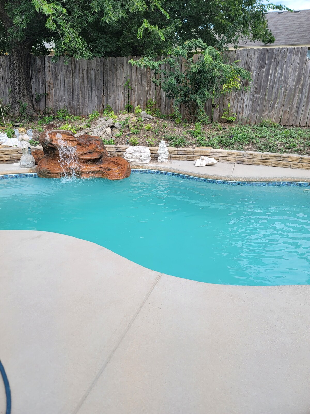 A serene pool is framed by natural stone and features a small rock waterfall. The clear water reflects the surrounding greenery and wooden fence. Decorative stone sculptures are placed around the pool area, adding a calming element to the outdoor space.