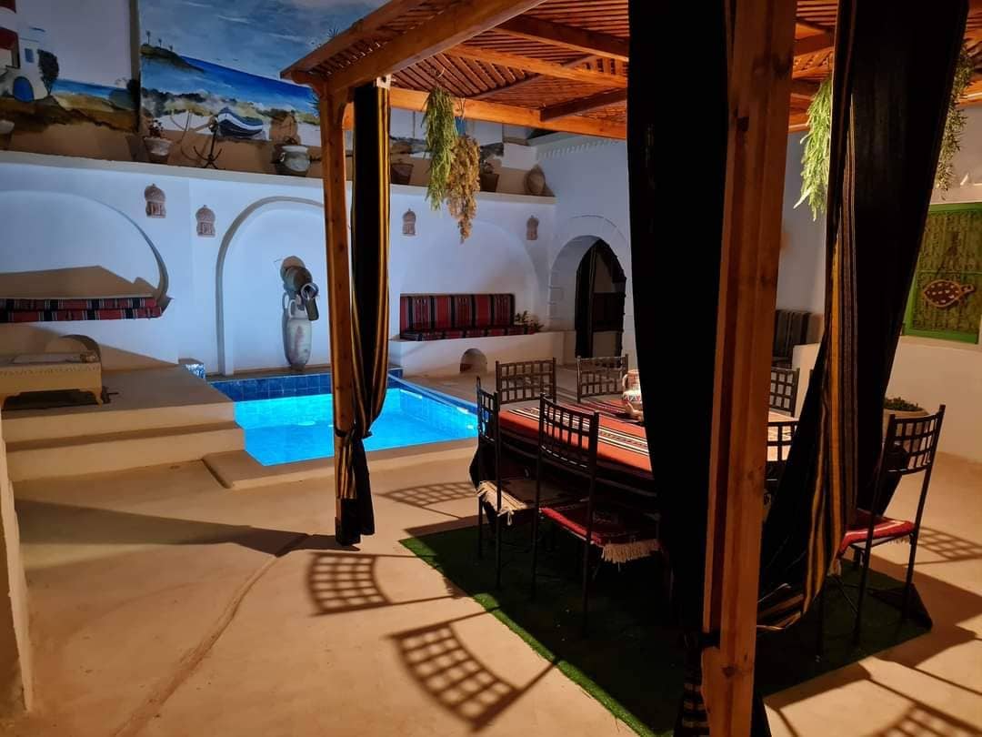 An indoor dining area is shown, featuring a large table surrounded by black metal chairs. The turquoise water of a private pool is visible nearby, under a wooden structure with decorative details. Natural light enters through a nearby opening, enhancing the space's inviting feel.