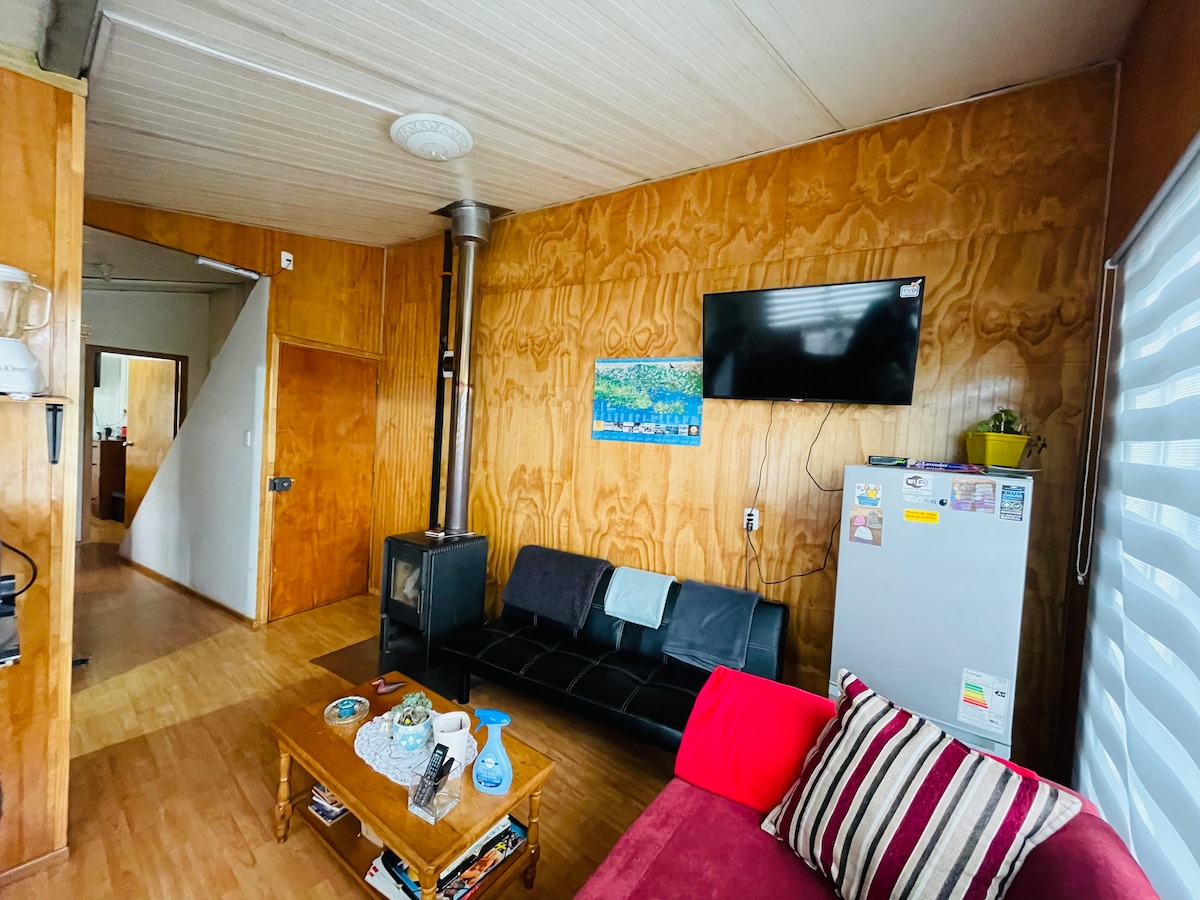 A living area features warm wooden walls and flooring, equipped with a black couch and a red upholstered chair. A wooden coffee table is positioned in front, displaying decorative items. A television is mounted on the wall, while a stove is visible nearby. The entry to the kitchen is observed in the background.