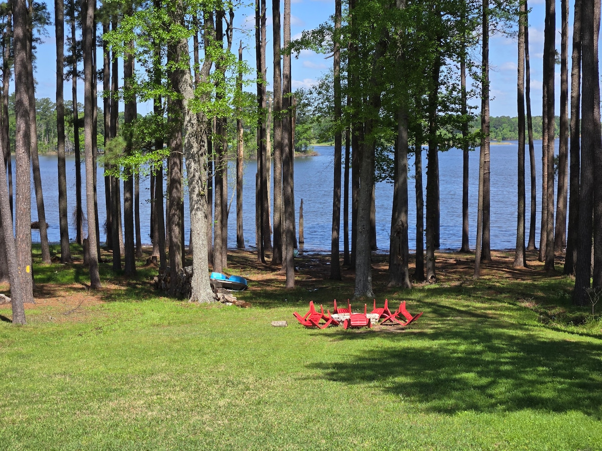 A serene lakeside setting is framed by tall, leafy trees. A vibrant, circular seating arrangement in red is placed on lush green grass, facing the tranquil water. The calm lake reflects the clear blue sky, contributing to a peaceful outdoor environment.