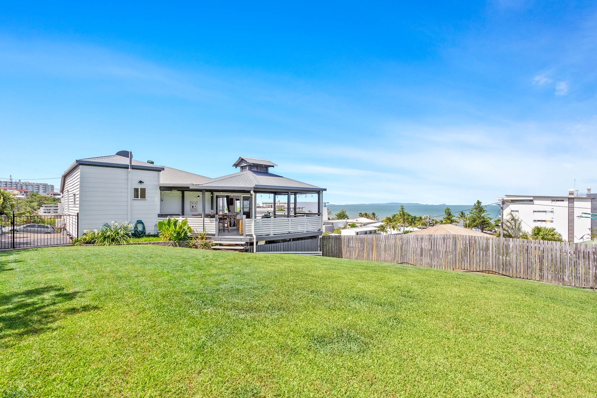 The exterior of the house is showcased, featuring a well-maintained grassy yard that is fully fenced. A spacious deck extends from the house, providing views of the ocean in the distance. The surrounding area includes nearby buildings and a clear blue sky.