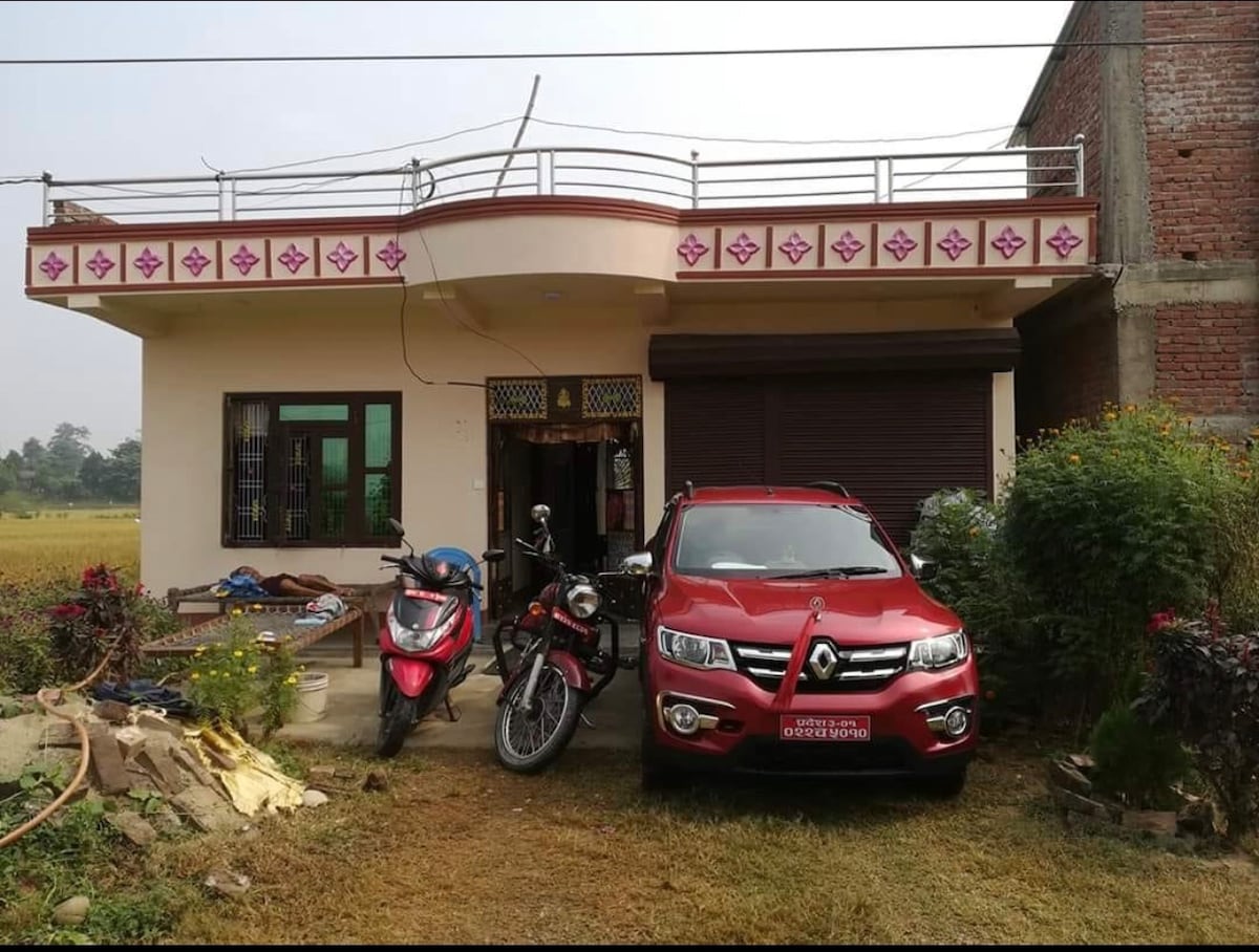 A charming exterior view of a single-story house is presented, featuring a welcoming entrance adorned with decorative elements. A red car and a motorcycle are positioned in front, with lush greenery and flowers surrounding the property, contributing to a tranquil outdoor setting.