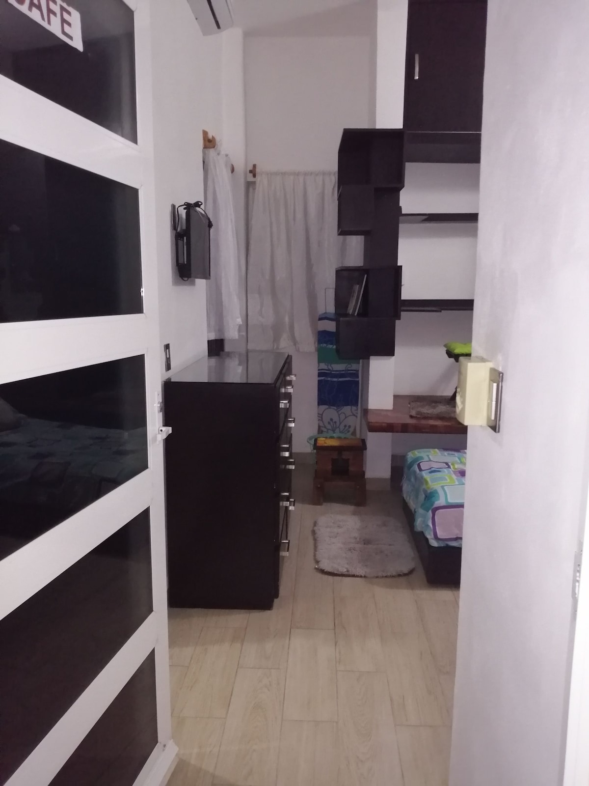 A hallway leads into a cozy room that features a charming dresser and shelf unit against the wall. The bed is neatly made with blue and patterned bedding. A small rug lies near the entrance, and sheer curtains frame the window, providing a touch of privacy.