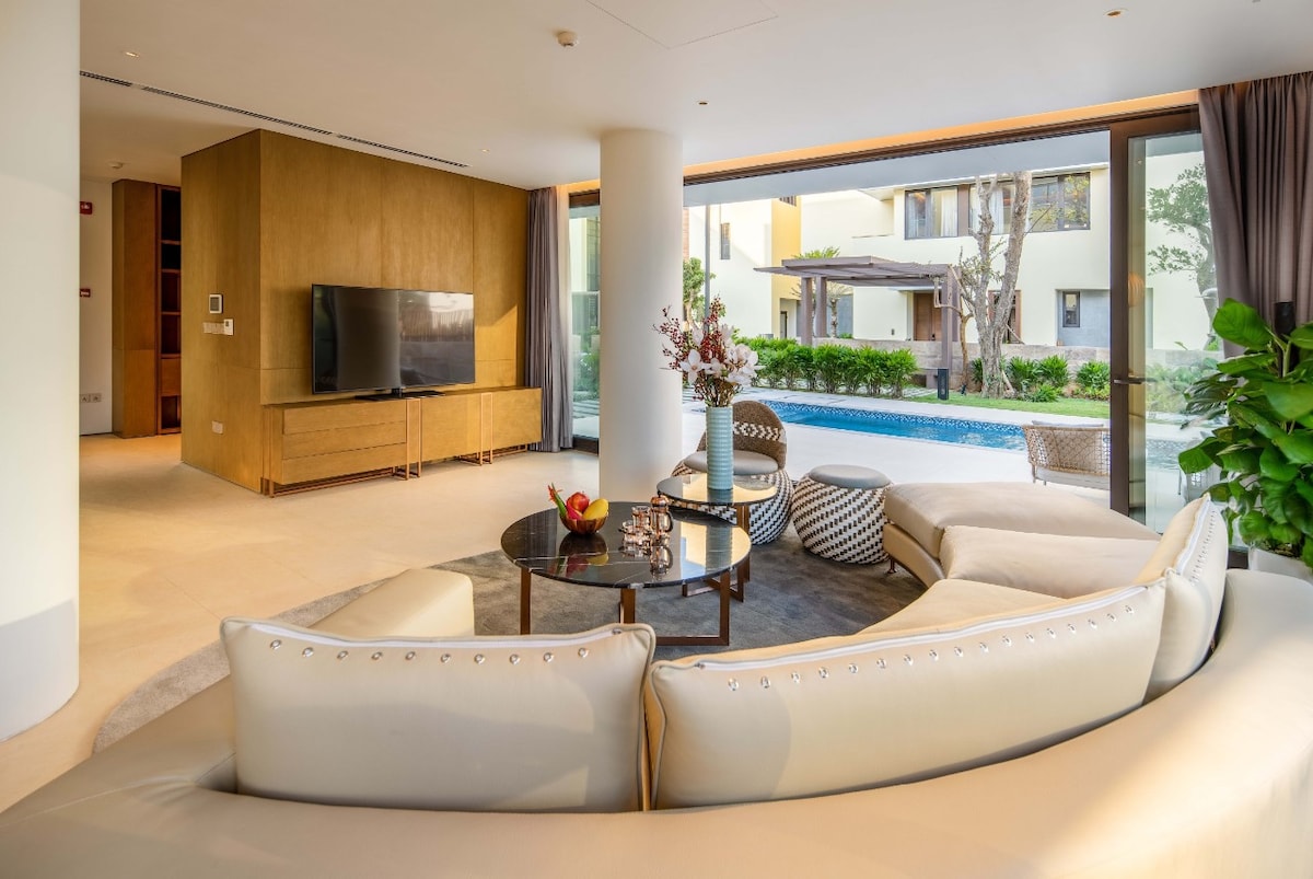 A modern living area features a circular white sectional sofa and a round glass coffee table, complemented by decorative elements. A large television is mounted on a wooden wall unit. Natural light floods the space through expansive glass windows, revealing a swimming pool and lush greenery outside.