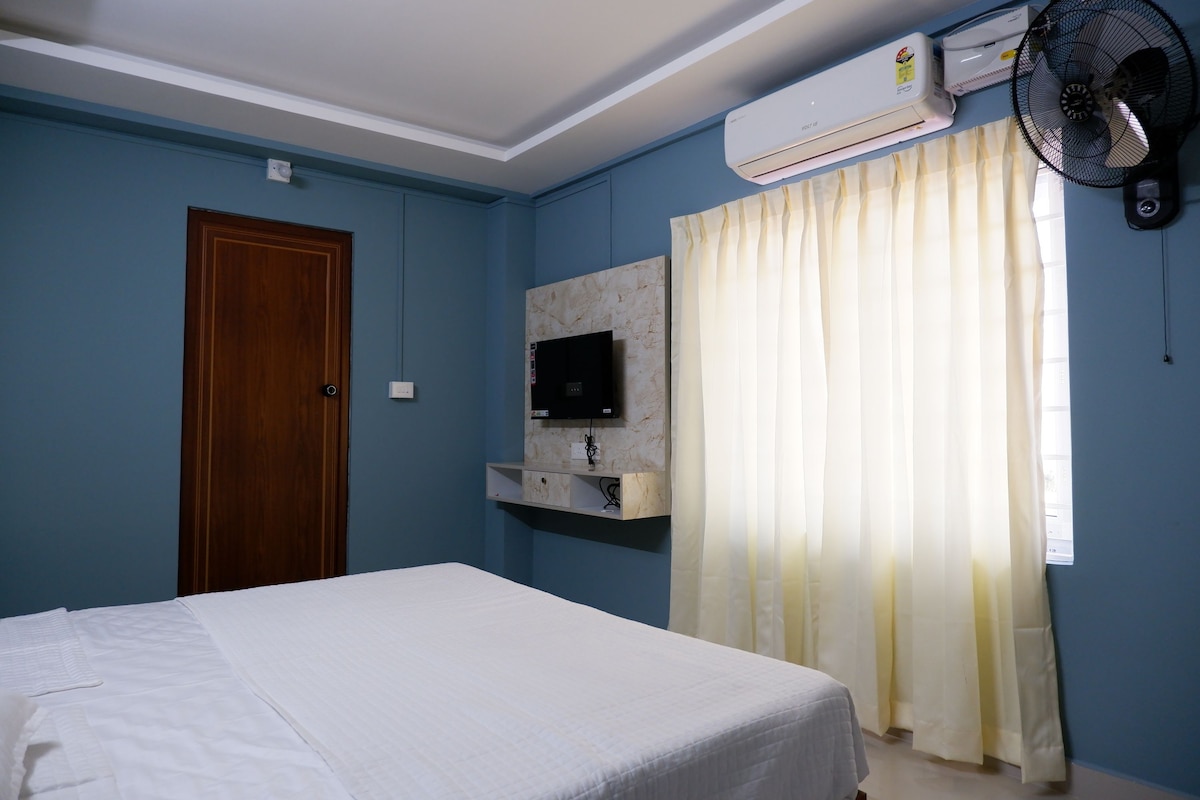 A well-lit room features a spacious bed draped in white linens. A wall-mounted television is set against a light-colored panel. Large windows dressed in sheer curtains allow natural light to brighten the blue walls, while a fan provides additional airflow.