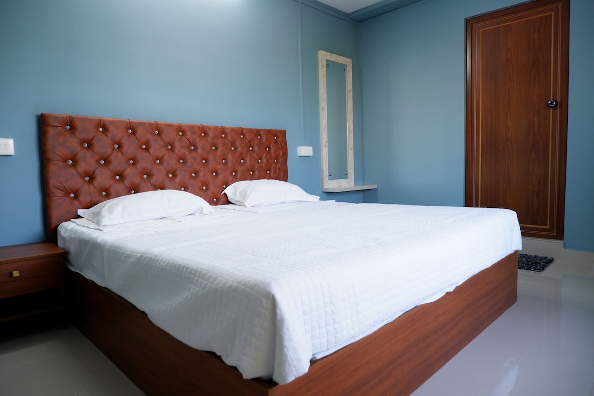 A spacious sleeping area features a tufted brown headboard with two white pillows atop a neatly made bed. Light blue walls complement the wooden accents, including a bedside table and a mirror hanging to the side. A door leads to another room.