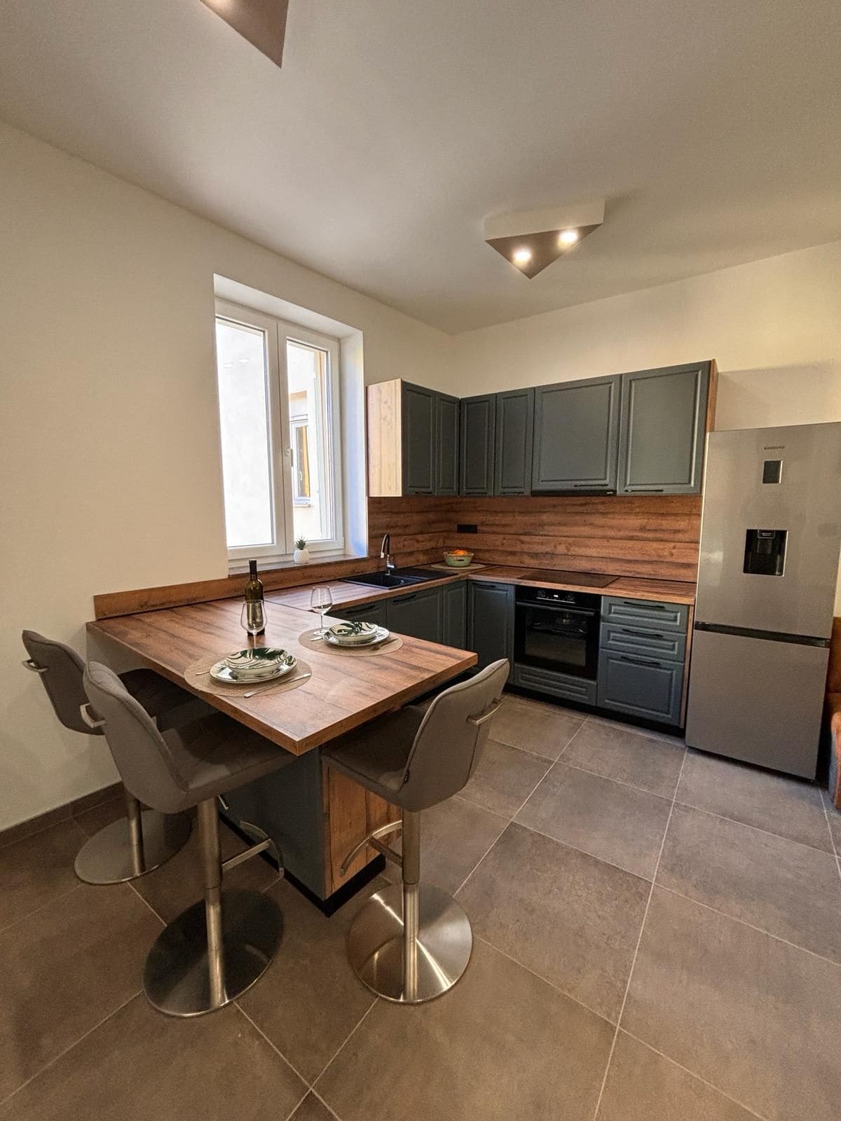 A modern kitchen showcases a blend of grey cabinets and wooden accents. A small dining space features two stools at a wooden counter. Stainless steel appliances are visible, complemented by a window that allows natural light to illuminate the area.