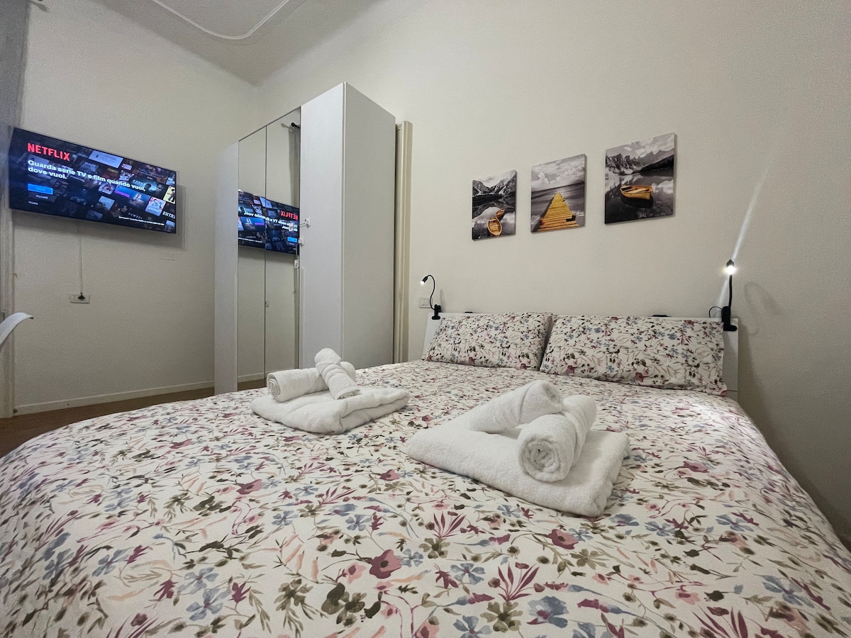 A bright sleeping area features a spacious bed with floral bedding and neatly arranged towels. A television mounted on the wall provides entertainment options, while a wardrobe is positioned nearby. Three framed landscape photographs add a decorative touch to the room.