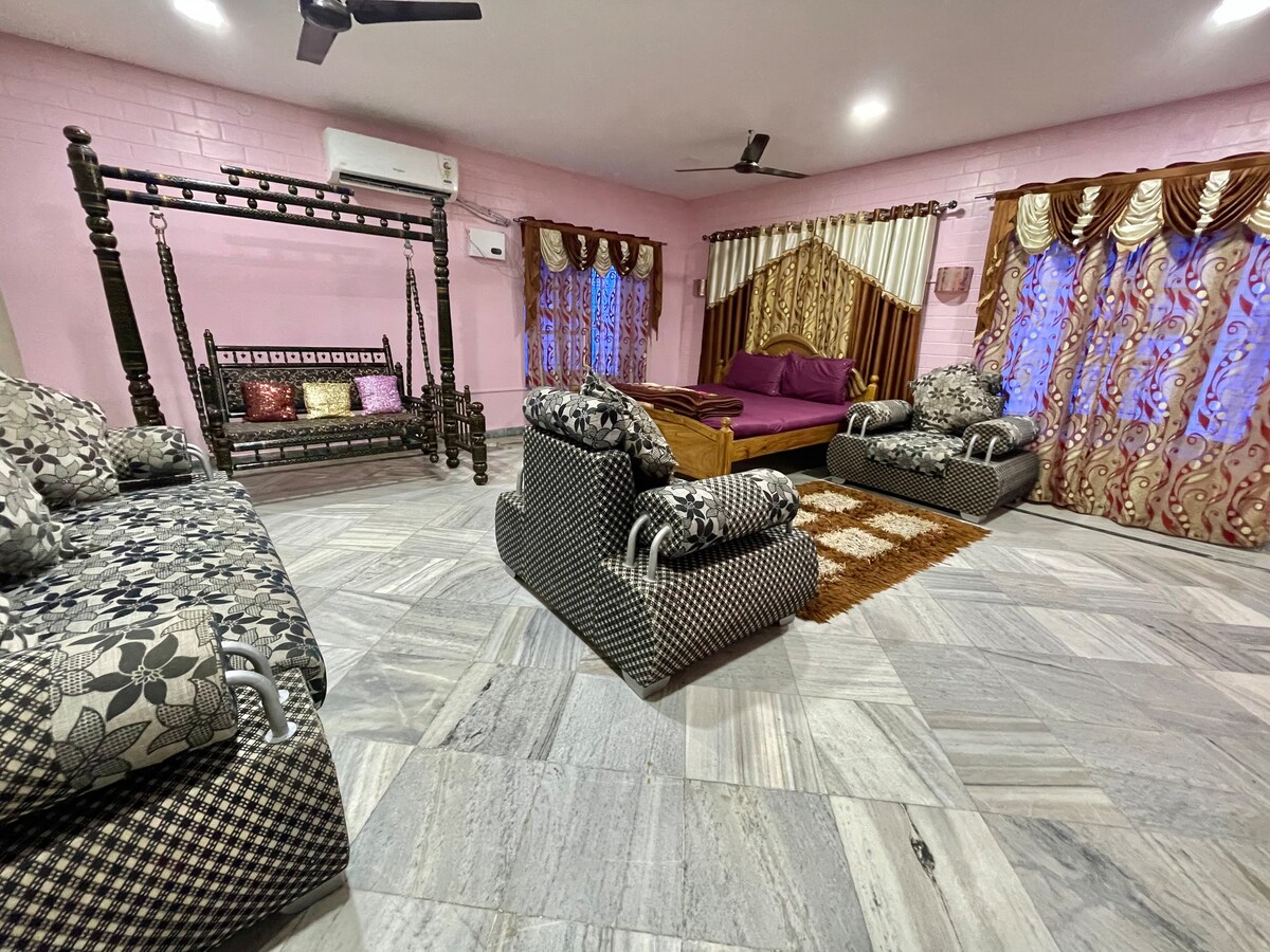 A spacious common room features a variety of seating arrangements, including patterned sofas and a traditional swing. Decorated curtains frame large windows, allowing light to illuminate the soft-colored walls. A light brown rug is positioned in the center, enhancing the room's inviting layout.