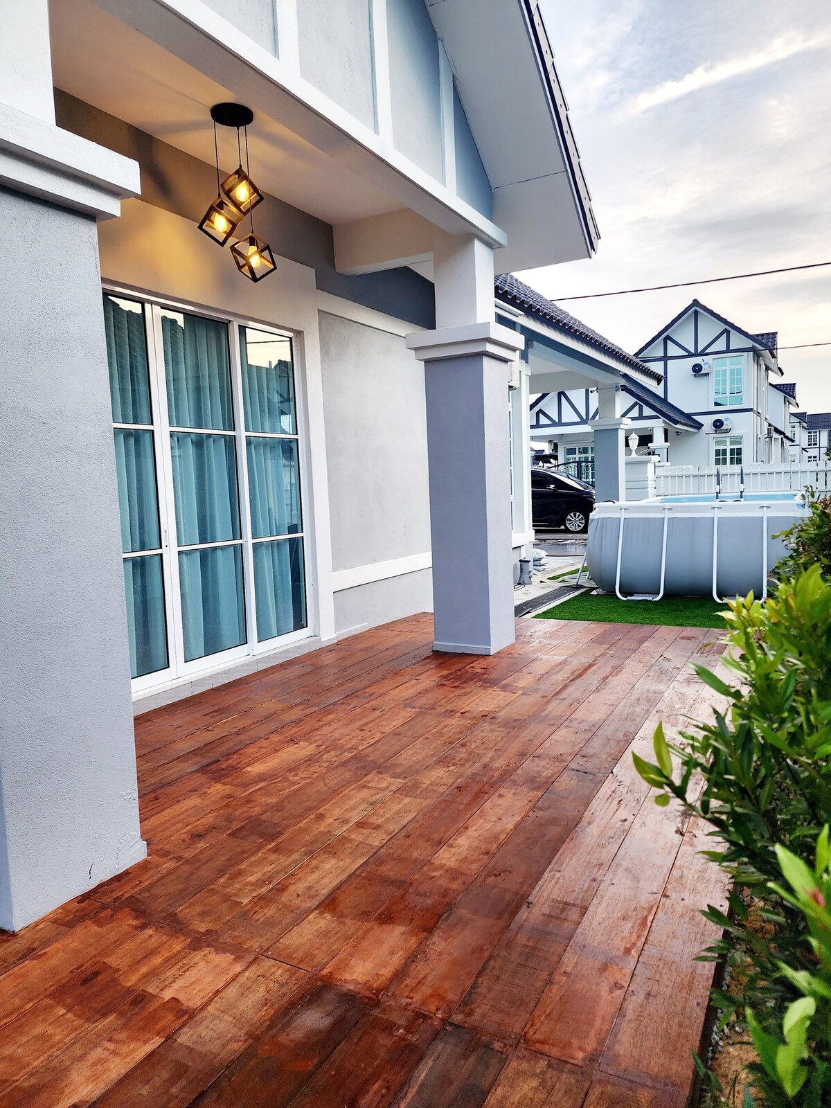 A wooden deck is presented in this image, featuring a smooth surface and ample space for relaxation. Large windows with light curtains are visible, allowing natural light to fill the interior. A swimming pool is situated nearby, framed by lush greenery.