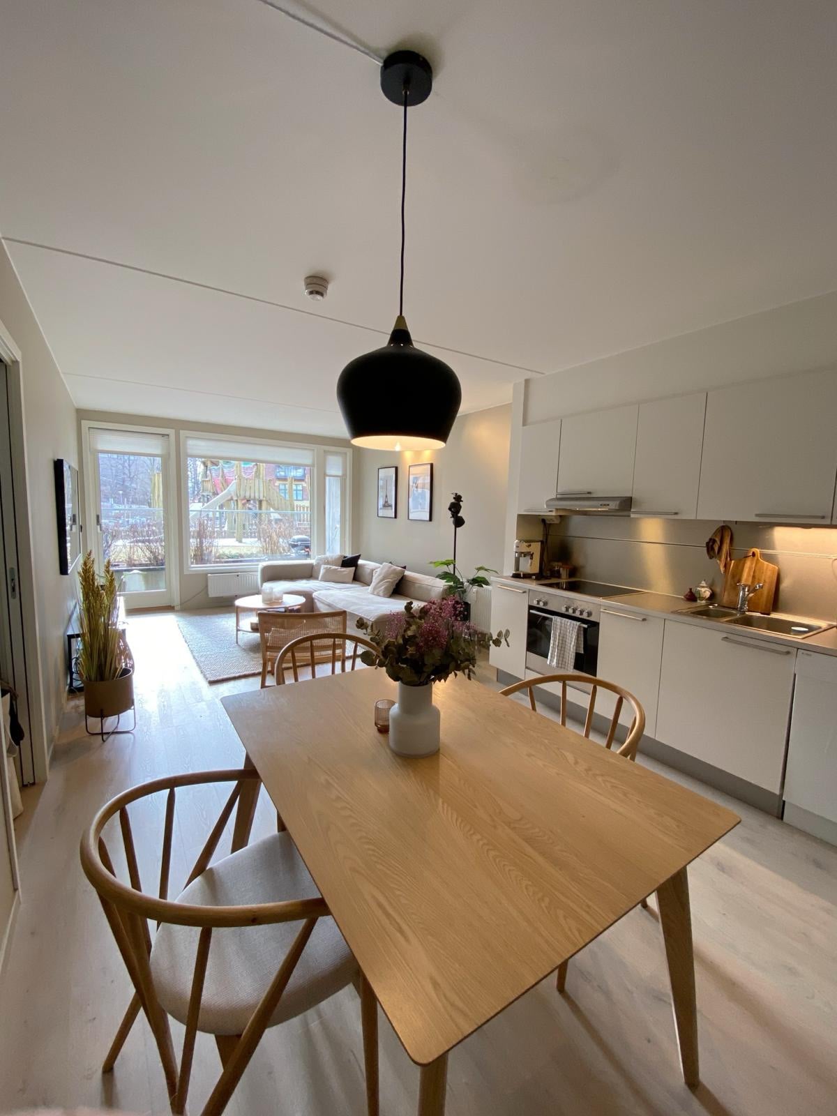 A modern dining area is highlighted by a wooden table surrounded by four chairs. A pendant light hangs above. In the background, a well-equipped kitchen is visible, along with a comfortable seating area featuring a large sofa and large windows allowing natural light into the space.