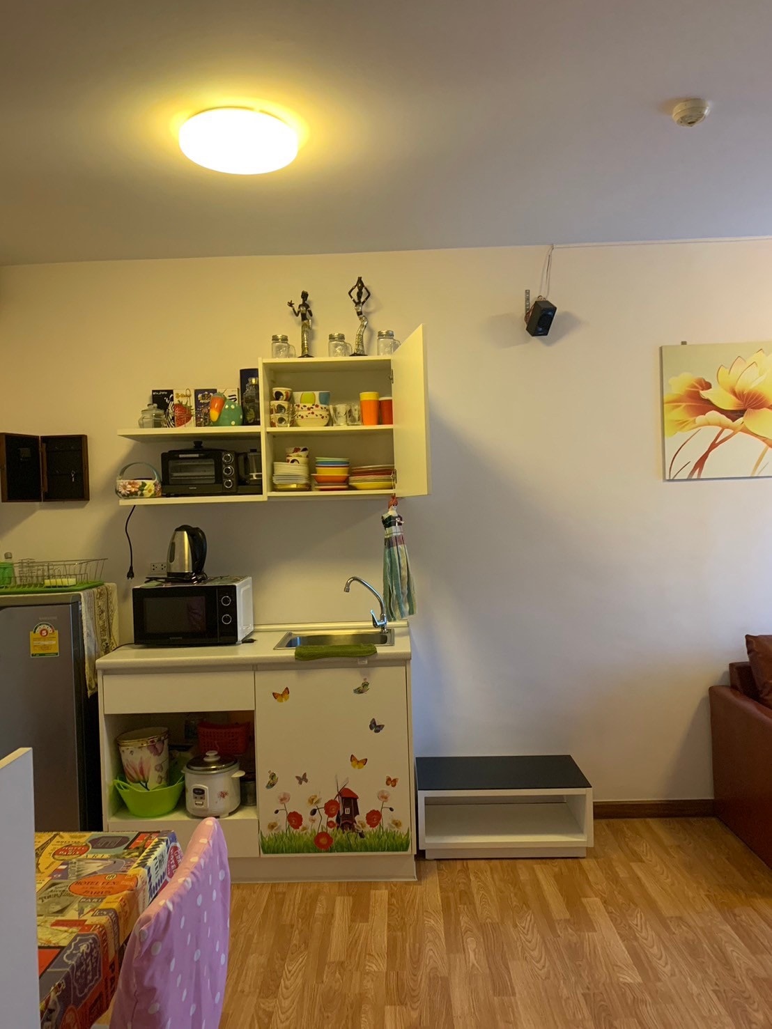 A compact kitchen area is visible, featuring white cabinets filled with colorful dishware. A microwave and kettle are placed atop a small countertop, beside a silver refrigerator. A wall-mounted speaker is noted, along with a floral artwork that adds a touch of decor to the space.