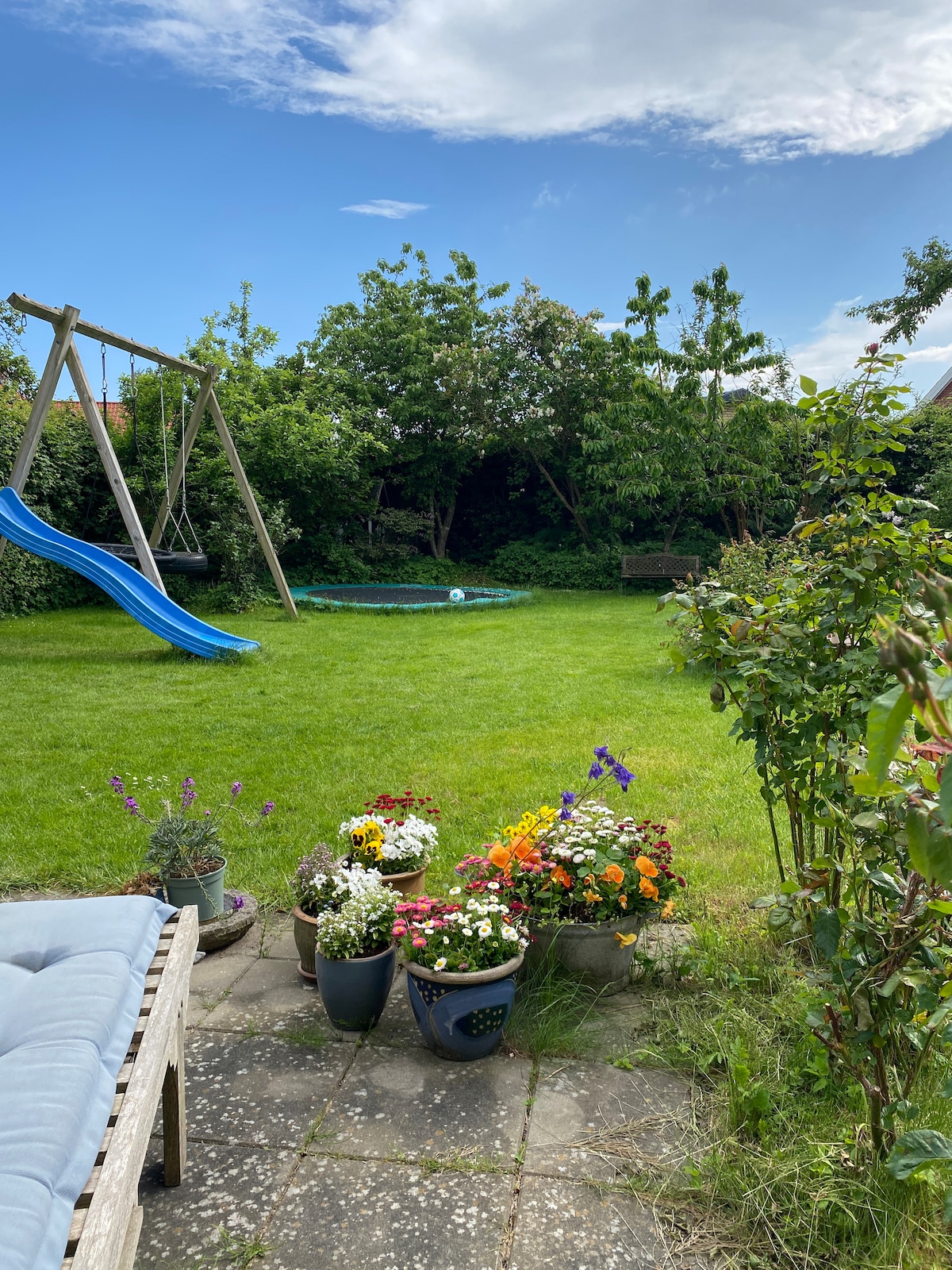 A spacious garden area is visible, featuring a sliding frame with a blue slide and a trampoline surrounded by lush greenery. Colorful flowerpots with blooming flowers are placed along the patio, adding a vibrant touch to the serene outdoor space under a clear blue sky.