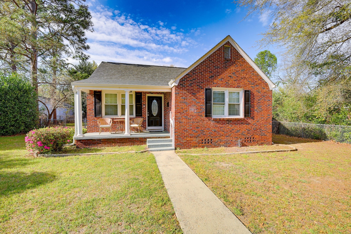 A single-story brick home is presented, featuring a welcoming front porch with seating options. Lush greenery surrounds the yard, and vibrant flowers add a pop of color. The entrance showcases a decorative door near large windows, providing a bright and inviting appearance.