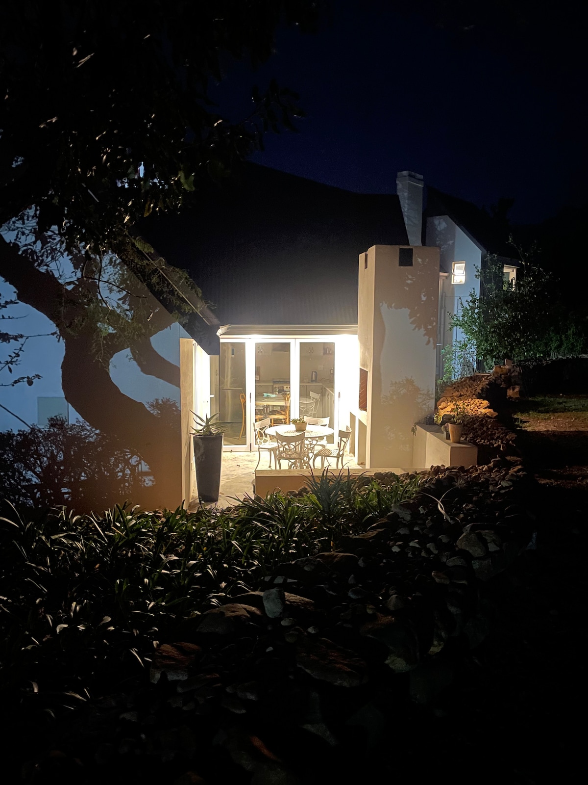 The exterior of the cottage is illuminated softly at night, with a light spilling from the large glass doors. Surrounding greenery frames a cozy outdoor seating area, while a sculpture enhances the peaceful setting among the trees and garden.