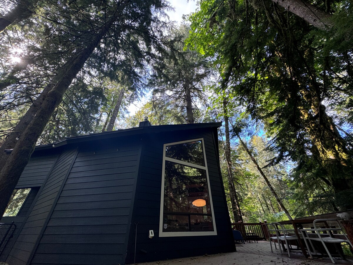 The cabin is nestled among towering trees, with large windows allowing natural light to fill the interior. A spacious deck extends from the cabin, offering a mix of seating options, while the surrounding forest creates a serene and secluded atmosphere.