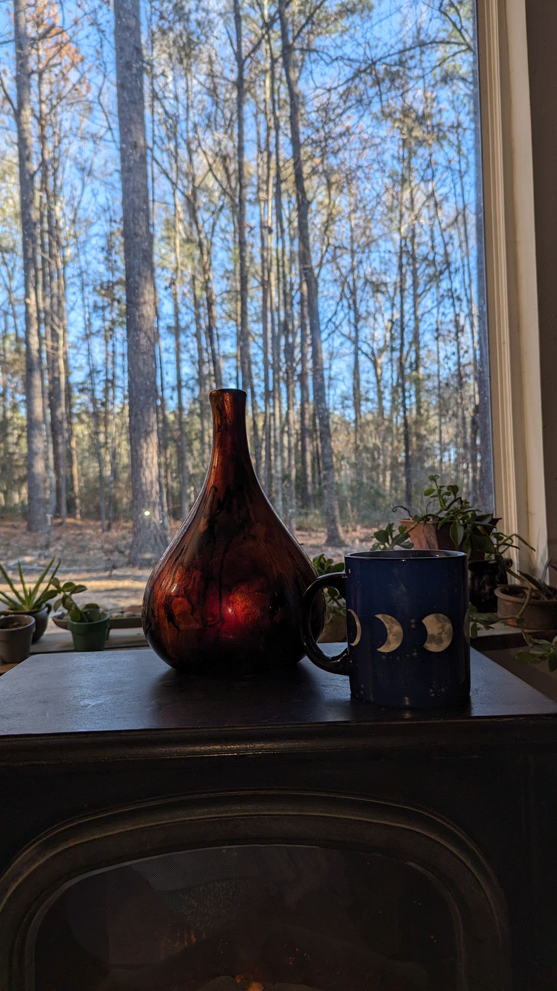 A decorative vase in a deep reddish-brown hue sits on a surface next to a mug adorned with moon phases. A large window reveals a backdrop of tall trees under a clear blue sky, enhancing the serene setting.