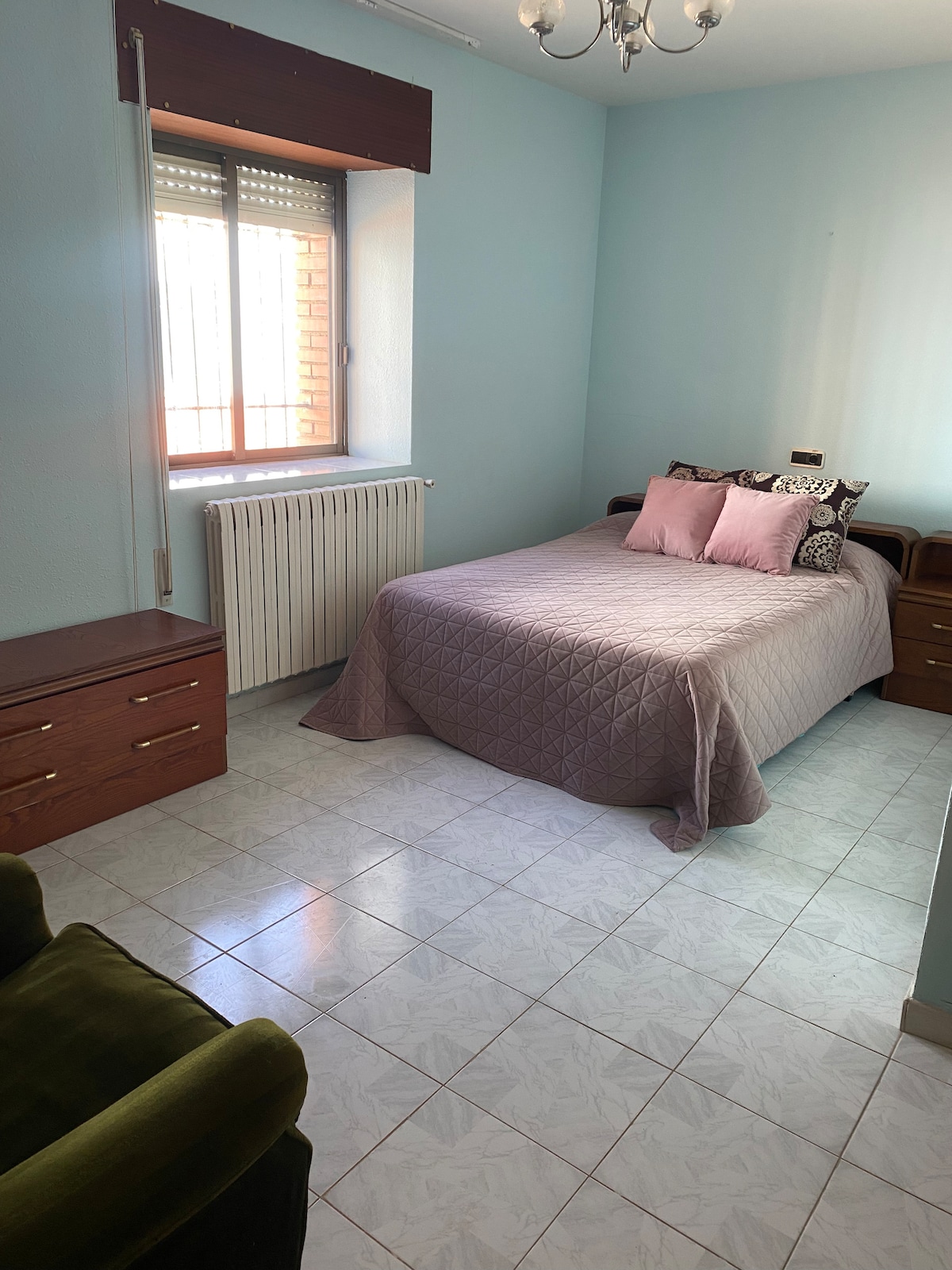 A serene bedroom features a spacious bed covered with a textured quilt. Two decorative pillows add subtle color. Natural light filters through a window dressed with sleek blinds, while a green chair and wooden nightstands complement the light blue walls.