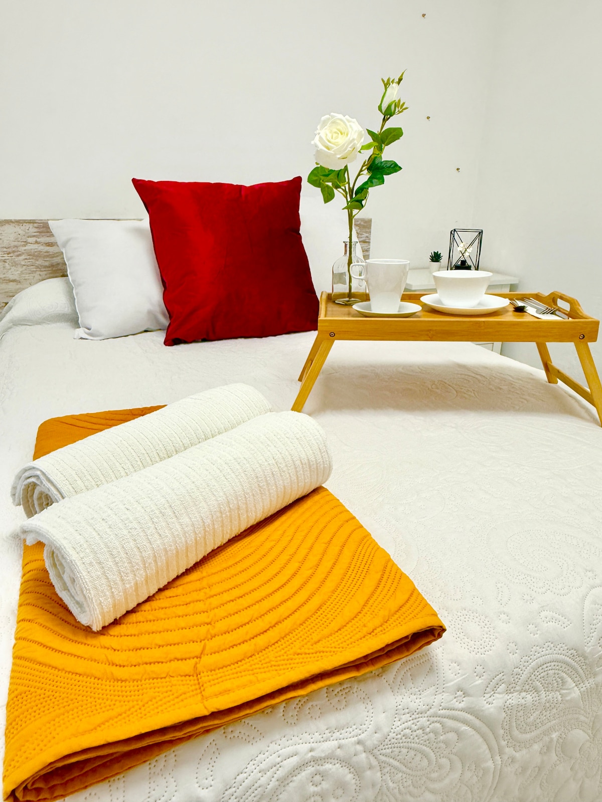 A cozy sleeping area features a spacious bed adorned with a white coverlet, complemented by decorative pillows in red and white. A tray with a bowl and cup is placed at the foot of the bed, alongside neatly rolled towels, creating an inviting atmosphere.
