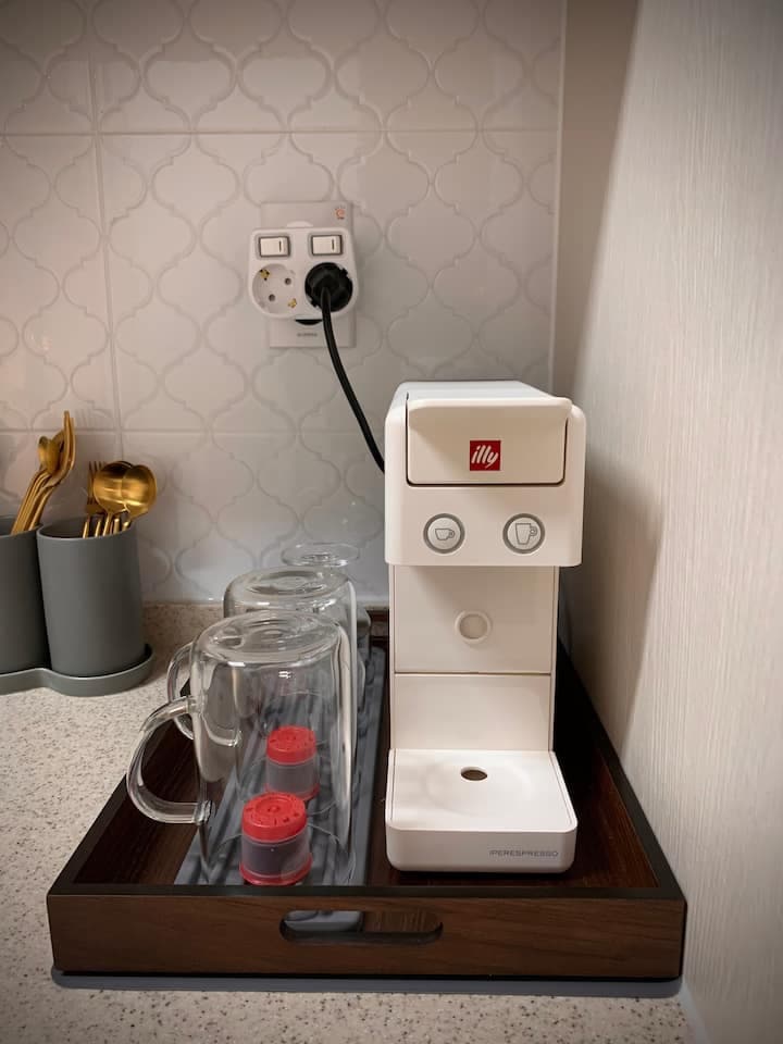 An Illy coffee machine is displayed on a wooden tray, accompanied by two glass mugs and a set of coffee capsules. The backdrop features a tiled wall with a power outlet, while golden cutlery rests in a gray holder beside the machine.