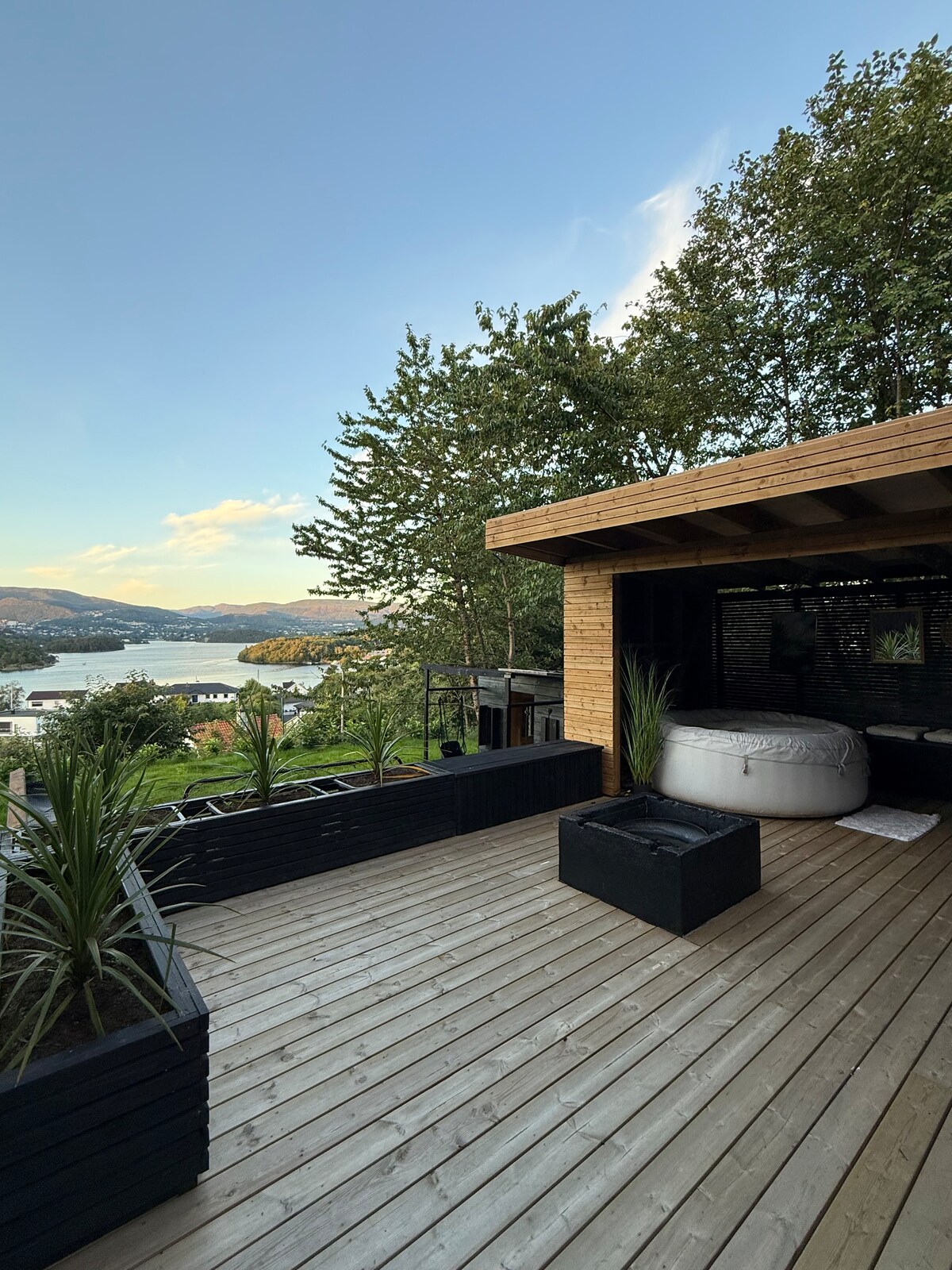 An outdoor terrace offers a view of the landscape, featuring a wooden deck and potted plants. A cozy seating area with a cushioned chair invites relaxation. The background reveals water and rolling hills, creating a serene environment.