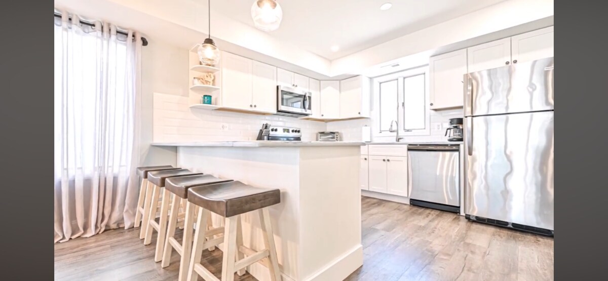 A modern kitchen features light cabinetry and stainless steel appliances. An island with four wooden bar stools provides a casual dining space, while natural light filters through a nearby window, complemented by sheer curtains.