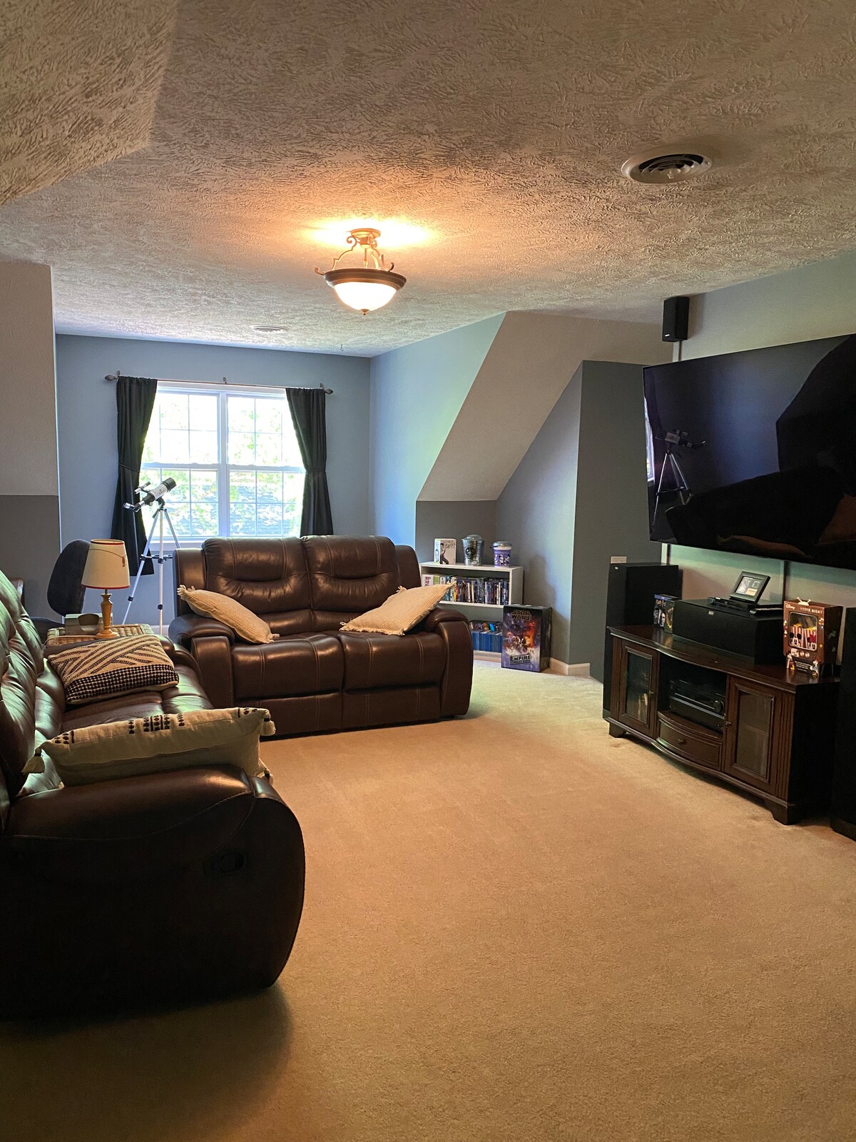 A cozy living room is highlighted by two brown recliner sofas facing a large flat-screen television. Soft carpet covers the floor, while large windows allow natural light to fill the space. A bookshelf is visible in one corner, enhancing the room's inviting ambiance.