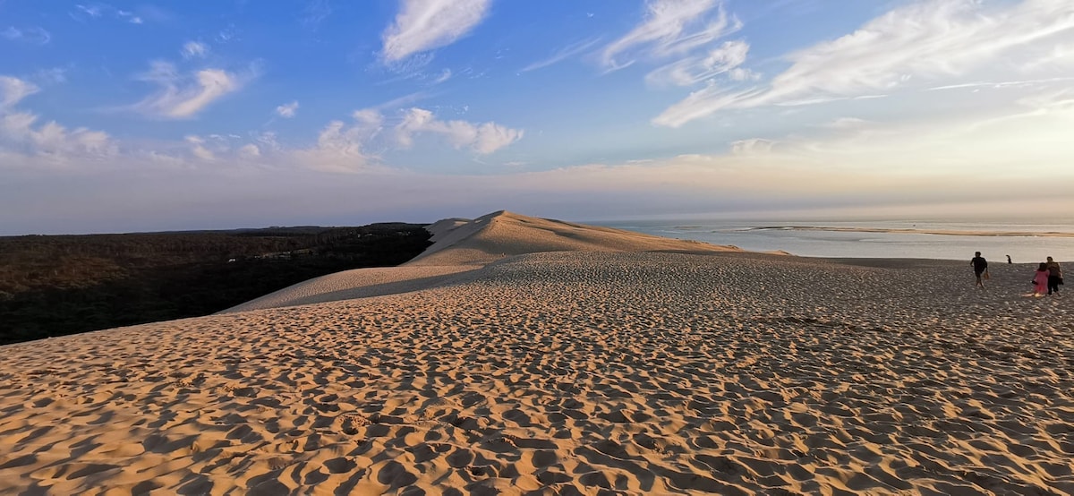Vast sandy dunes stretch toward the horizon, illuminated by a pastel sky at sunset. Footprints mark the soft sand, while a distant pair of figures can be seen walking along the ridge, suggesting a peaceful exploration of this natural landscape.