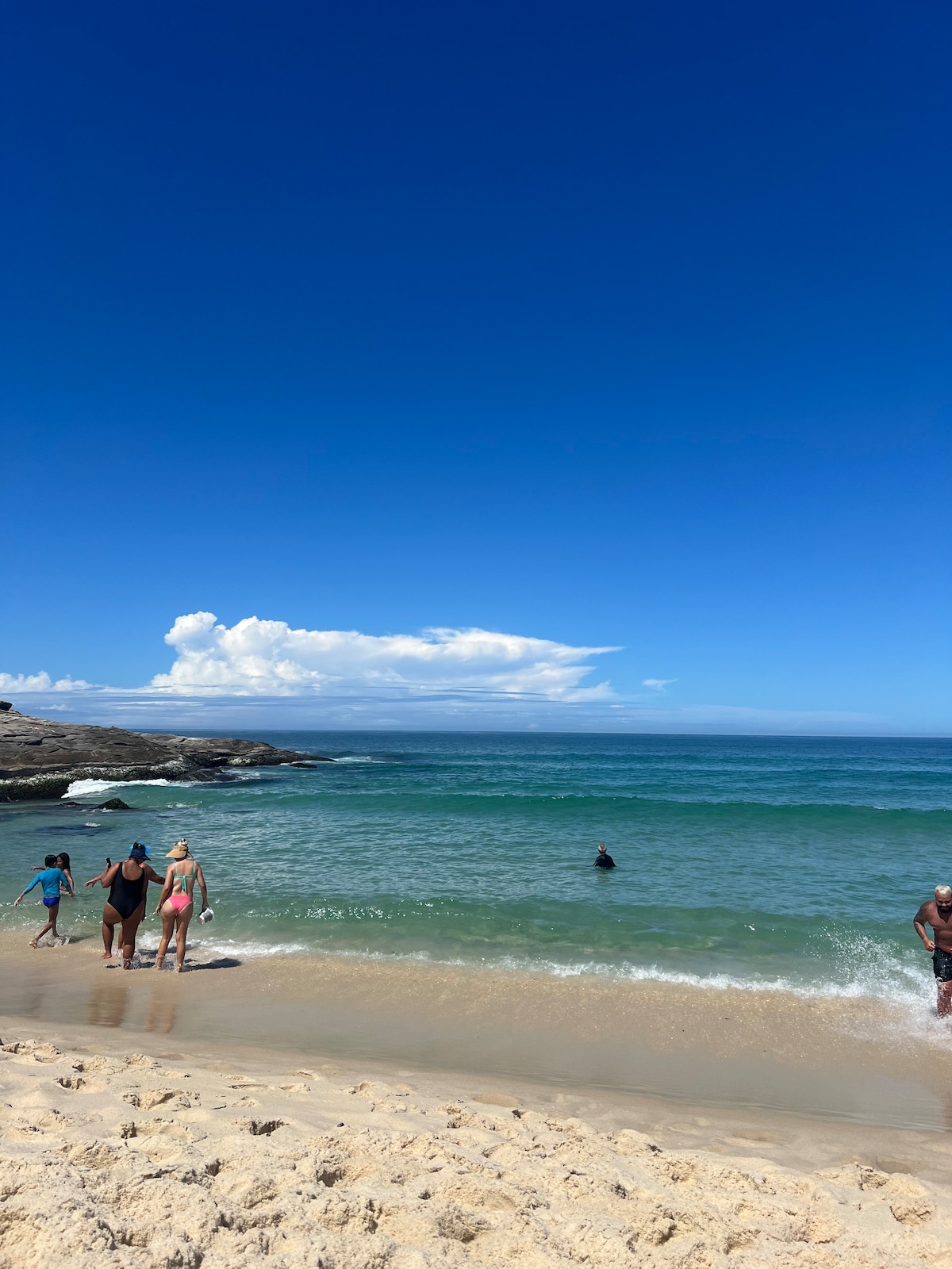 A sandy beach is depicted with gentle waves lapping at the shore. A clear blue sky dominates above, while a few clouds are visible in the distance. Several individuals are seen enjoying the water, highlighting a lively and inviting beach atmosphere.