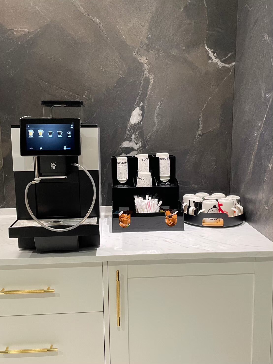 A sleek coffee machine with a touchscreen display is positioned atop a white marble countertop. Adjacent, a black organizer holds disposable straws and cups, providing convenient access. The backdrop features elegant dark stone walls, enhancing the modern feel of the coffee station.