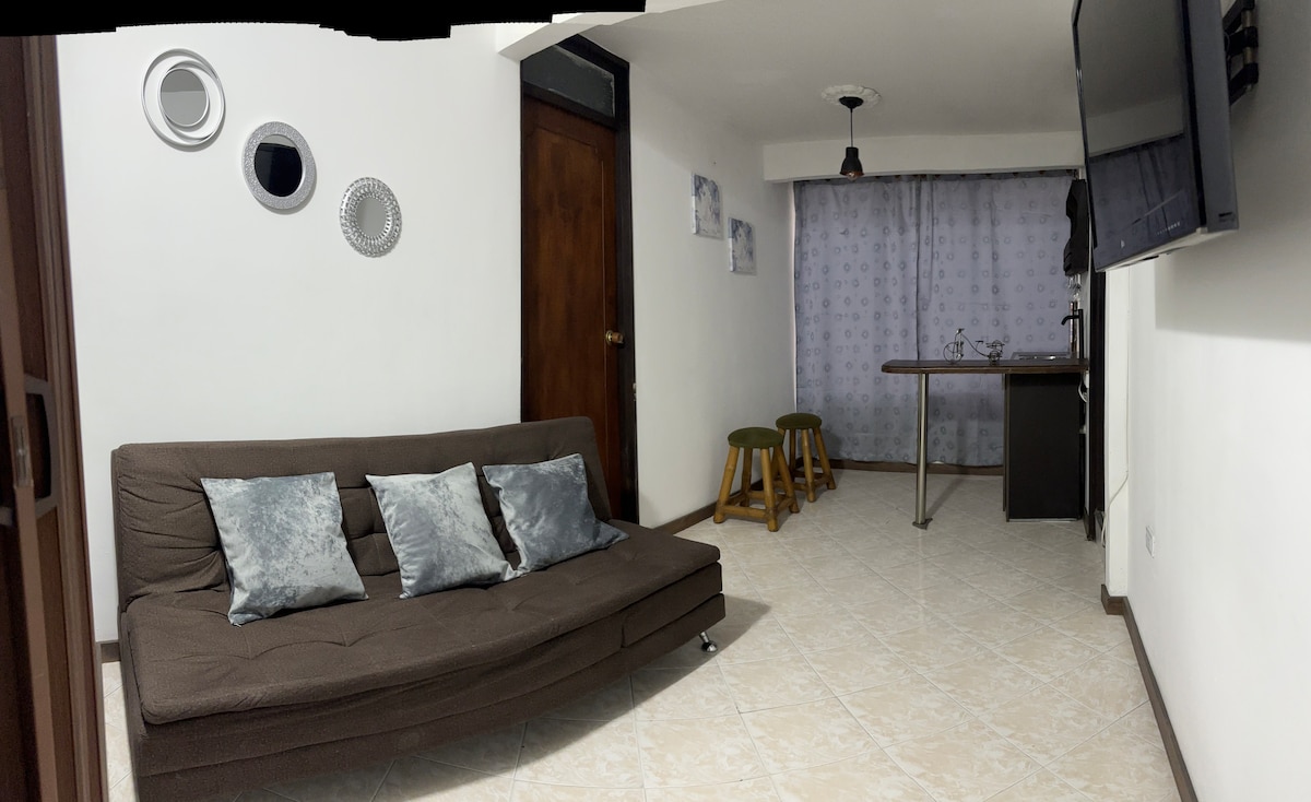 A spacious living area is highlighted by a comfortable sofa bed with decorative cushions. Two bar stools are positioned at a counter, with a television mounted on the wall. Soft lighting is provided by a pendant fixture, while a curtain softly drapes in the background.