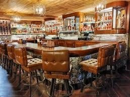 The image captures an elegantly designed bar area, featuring a curved wooden counter with plush leather stools. Shelves lined with a variety of spirits illuminate the space. Warm lighting from chandeliers enhances the inviting ambiance, reflecting a detail-oriented design with rich wood finishes.