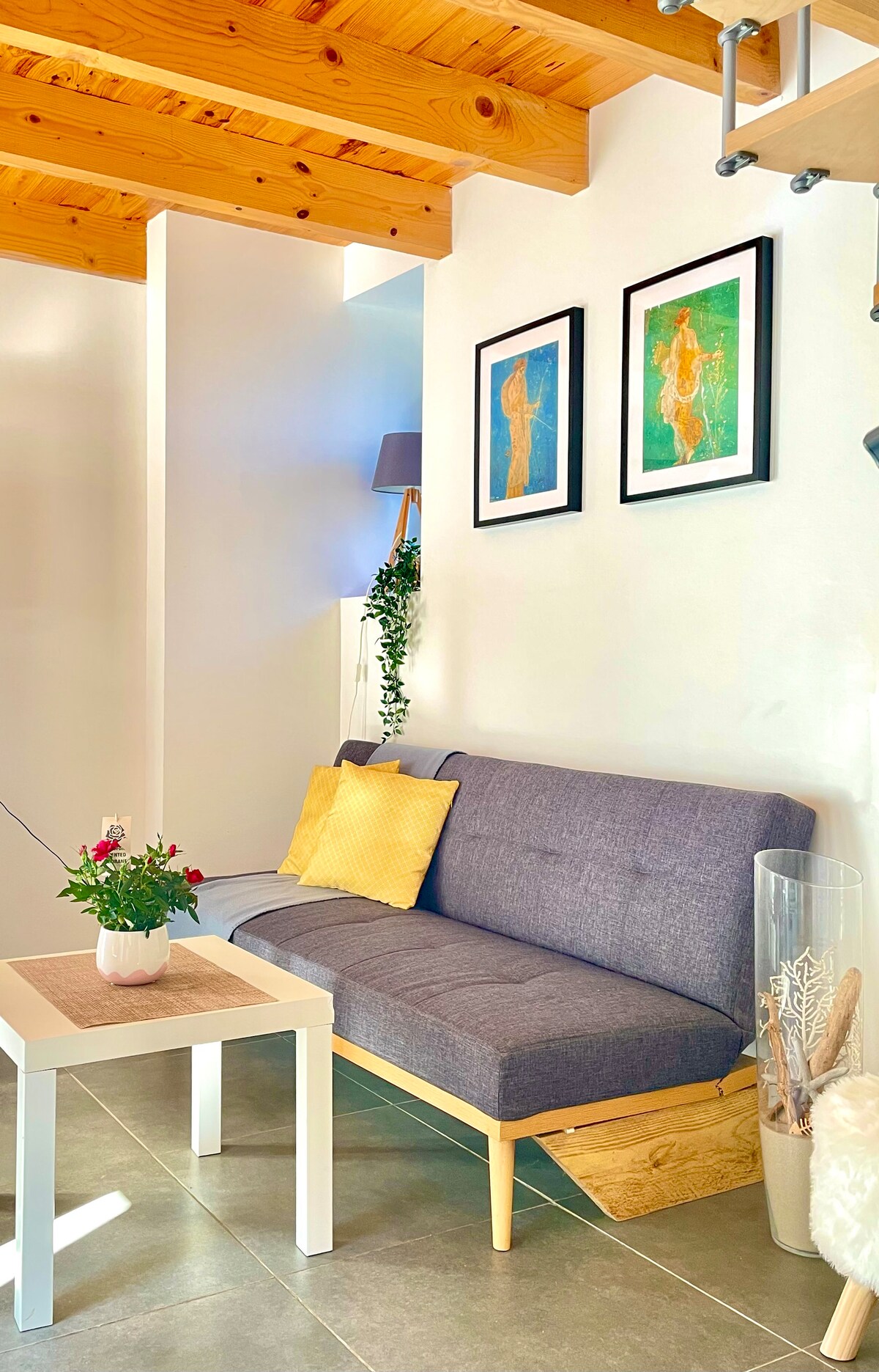 A cozy living area is displayed, featuring a grey sofa with cushions and a small white table. Two framed artworks are mounted on the wall above the sofa. Natural light filters in, illuminating the light-colored walls and wooden ceiling beams.